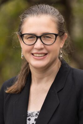 Portrait of Kirsten Turlo, a lecturer in the UCLA Biomedical Research Minor, wearing glasses and a black blazer over a patterned top, smiling outdoors against a blurred natural background.