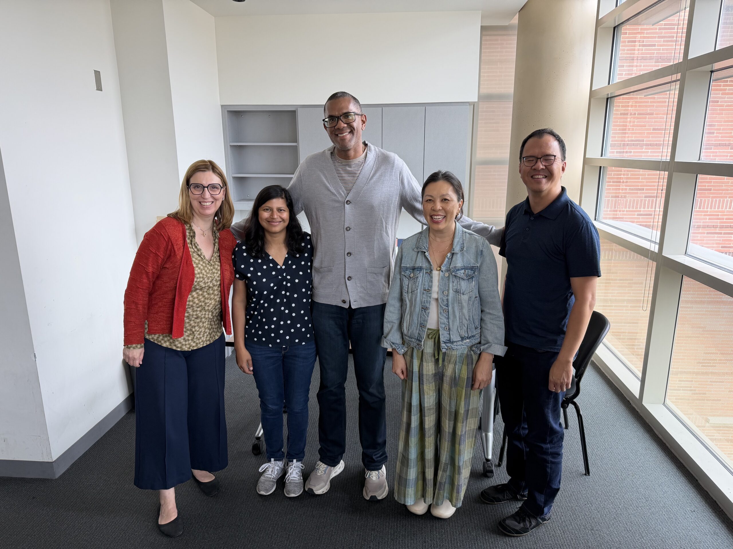 Five InDialogue past fellows stand side by side, smiling together in a brightly lit office space next to large windows.