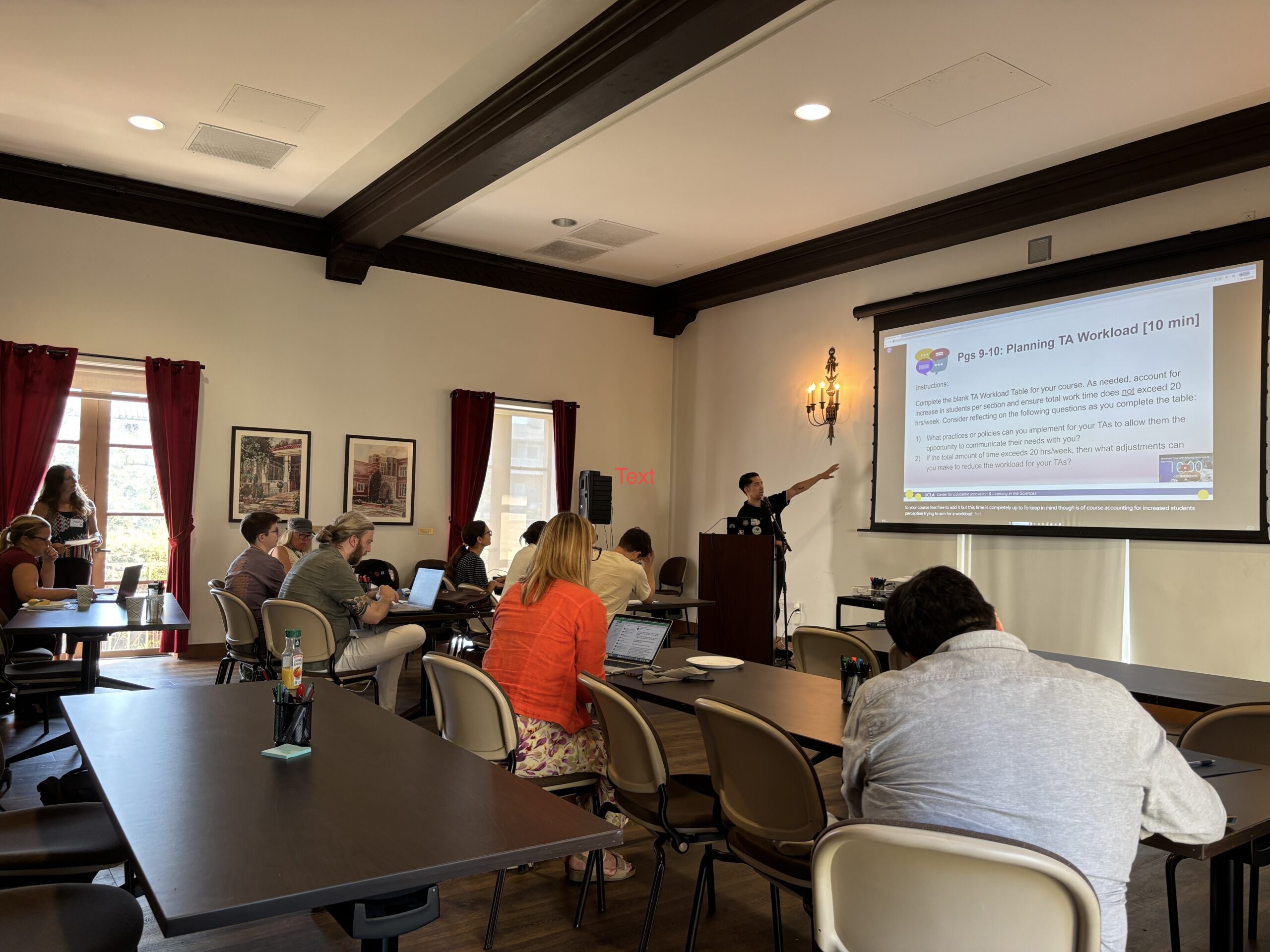 Professional development workshop with participants seated at tables using laptops and taking notes while a presenter points to a projected slide about planning teaching assistant workloads.

