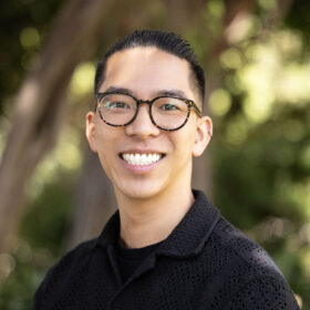 Portrait of a smiling man with short dark hair, round glasses, and a black collared shirt, facing the camera in an outdoor setting. 