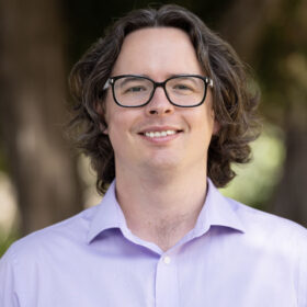Portrait of a smiling man with with medium-length wavy brown hair, rectangular glasses, and a light purple collared shirt, facing the camera in an outdoor setting.