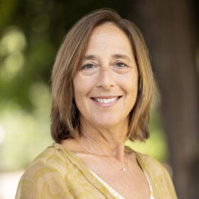 Portrait of a smiling woman with shoulder-length light brown hair, wearing a yellow patterned top and delicate necklace, facing the camera in an outdoor setting.