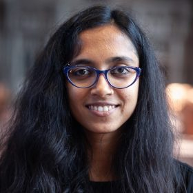 Portrait of a smiling woman with long, dark hair, wearing blue-framed glasses and dark top, facing the camera