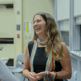 Portrait of a smiling woman with long wavy brown hair and glasses, wearing a black sleeveless top, colorful scarf, and bracelets, standing indoors.